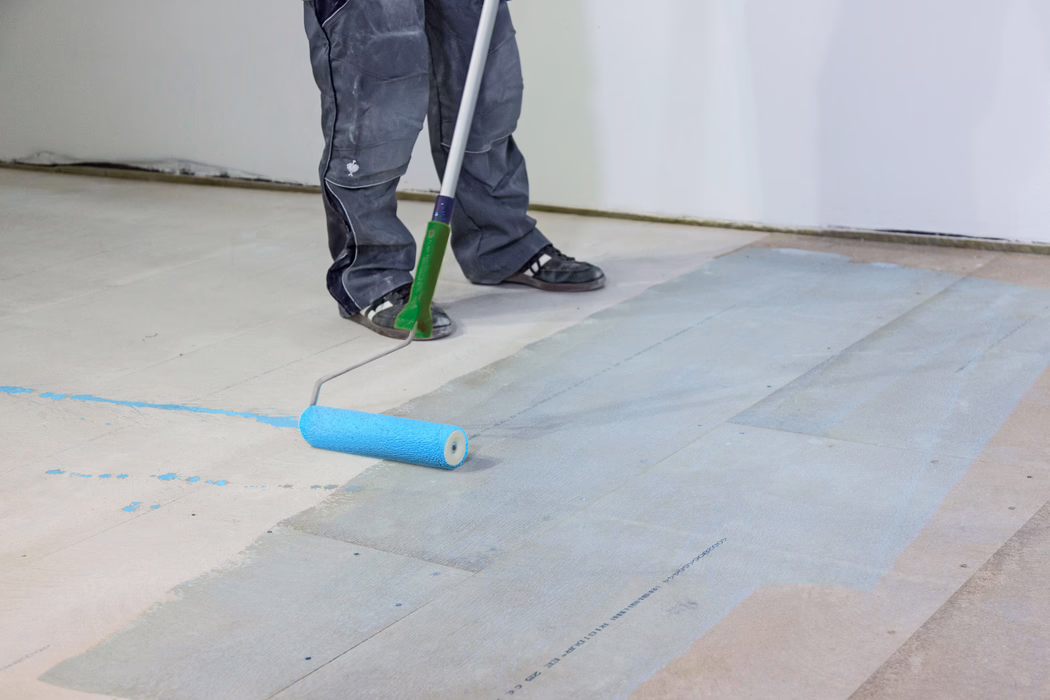 On voit les jambes et les pieds inférieurs d'un ouvrier qui applique un primaire ou une peinture bleu clair sur un sous-plancher en bois à l'aide d'un rouleau à long manche. La personne porte un pantalon de travail foncé et des baskets noires ; le rouleau laisse un film bleu humide sur les lames de plancher dans une pièce intérieure aux murs blancs. L'image met l'accent sur la préparation du sol, les outils et les éclaboussures de peinture.