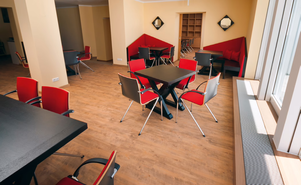Un espace lounge ou salle à manger moderne et lumineux avec un sol en bois clair, des murs beiges, des tables carrées noires et des chaises rouges rembourrées aux pieds chromés. De grandes fenêtres à droite laissent entrer la lumière naturelle ; des banquettes d'angle rouges intégrées, des étagères en arrière-plan et une grille de radiateur le long de la fenêtre complètent l'aménagement soigné, adapté aux réunions informelles ou aux repas.