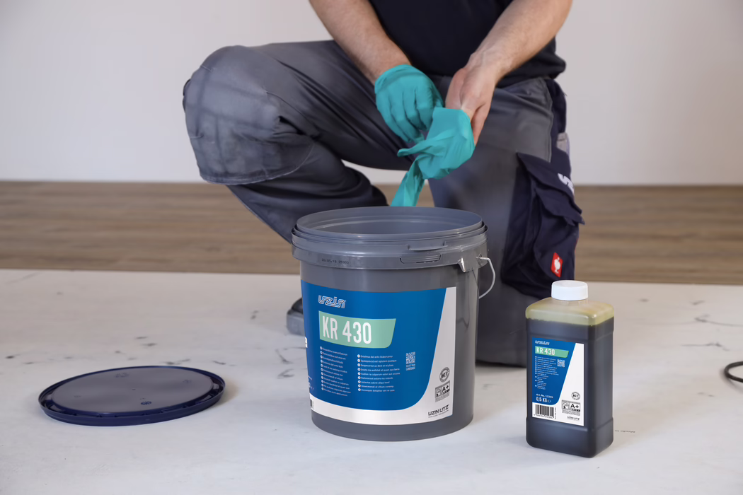 UZIN_Application_Image_KR 430_2019-06 Une personne à genoux met des gants jetables turquoise à côté d'un seau gris étiqueté 'KR 430' et d'une petite bouteille de liquide.