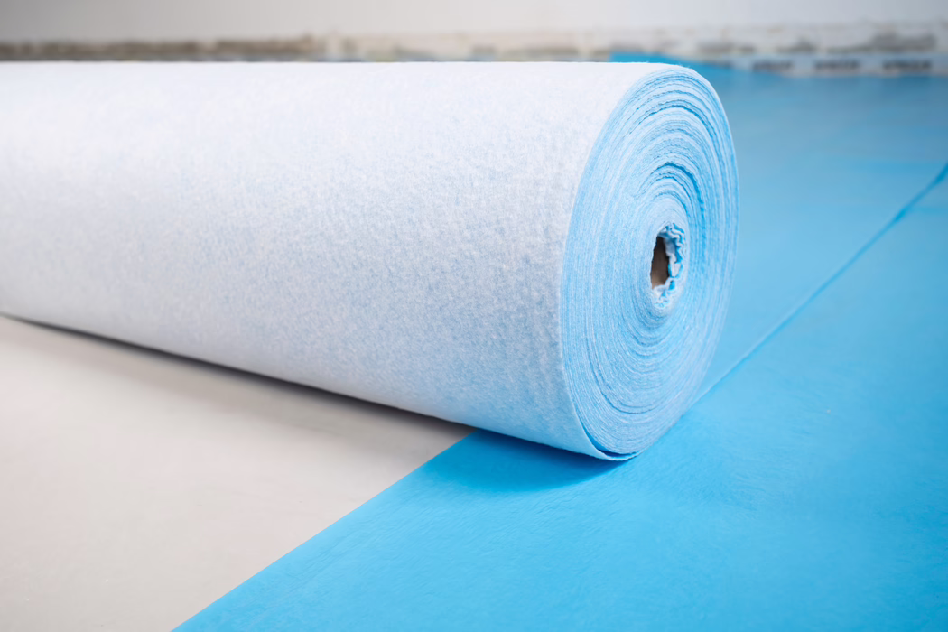 UZIN_Supplements & Accessories_PFB 160_blue_2024-06 (9) Gros plan d'un grand rouleau de tissu non tissé bleu clair ou de sous-couche posé sur une feuille bleue assortie. L'image montre la surface texturée et fibreuse et les couches en spirale enroulées autour d'un noyau en carton, posé sur un sol clair. Un éclairage doux et diffus met en valeur les fibres subtiles et la forme cylindrique ; l'arrière-plan est flou.