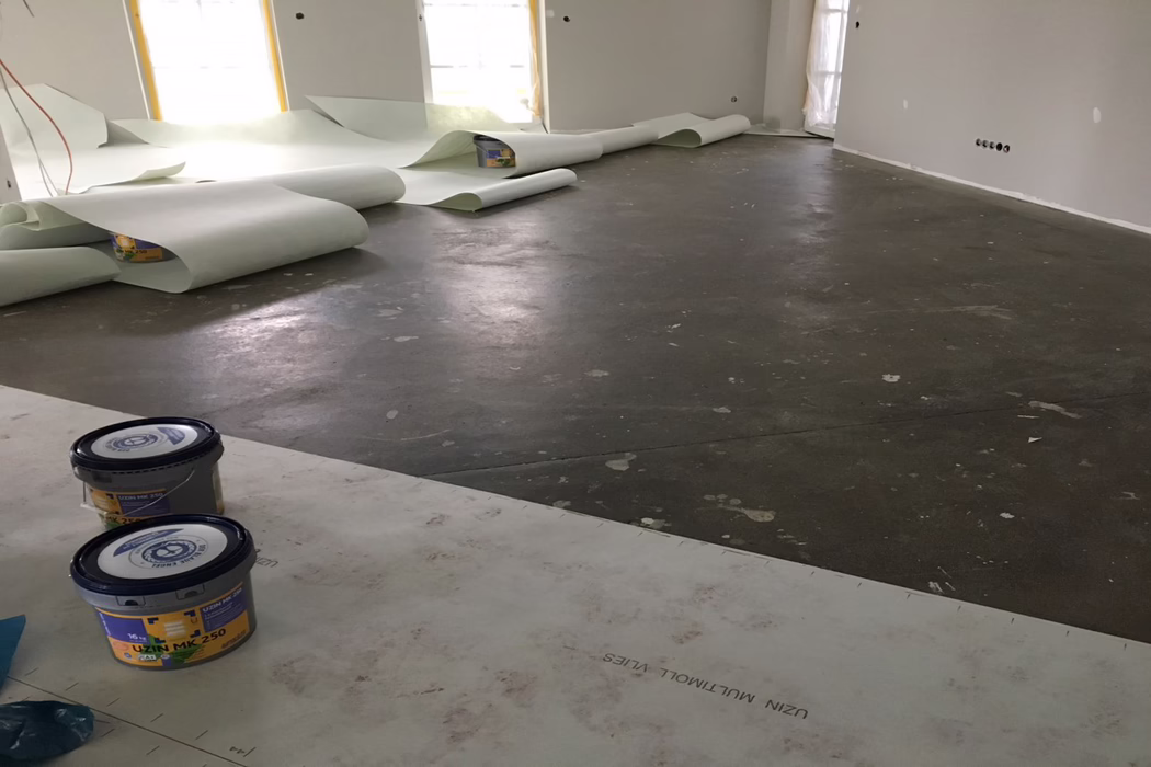 UZIN_Application_Image_Multimoll Vlies_2019-06 Intérieur d'une pièce d'habitation en cours de rénovation : un grand espace ouvert avec une chape en béton brute et sombre, marquée de projections de peinture, d'empreintes et de zones réparées. Le long du mur du fond, plusieurs larges rouleaux de sous-couche ou de membrane de sol vert pâle sont partiellement déroulés près de fenêtres protégées par un film. Au premier plan, deux seaux en plastique de colle ou d'apprêt (marqués UZIN MK 250) reposent sur des panneaux de protection ; les murs sont fraîchement enduits ou apprêtés en gris clair et laissent apparaître des boîtiers électriques et des prises exposées. Une lumière naturelle diffuse filtre à travers les fenêtres couvertes et crée de doux reflets sur le sol ; aucun ouvrier ni outil n'est visible, indiquant une étape d'installation interrompue.