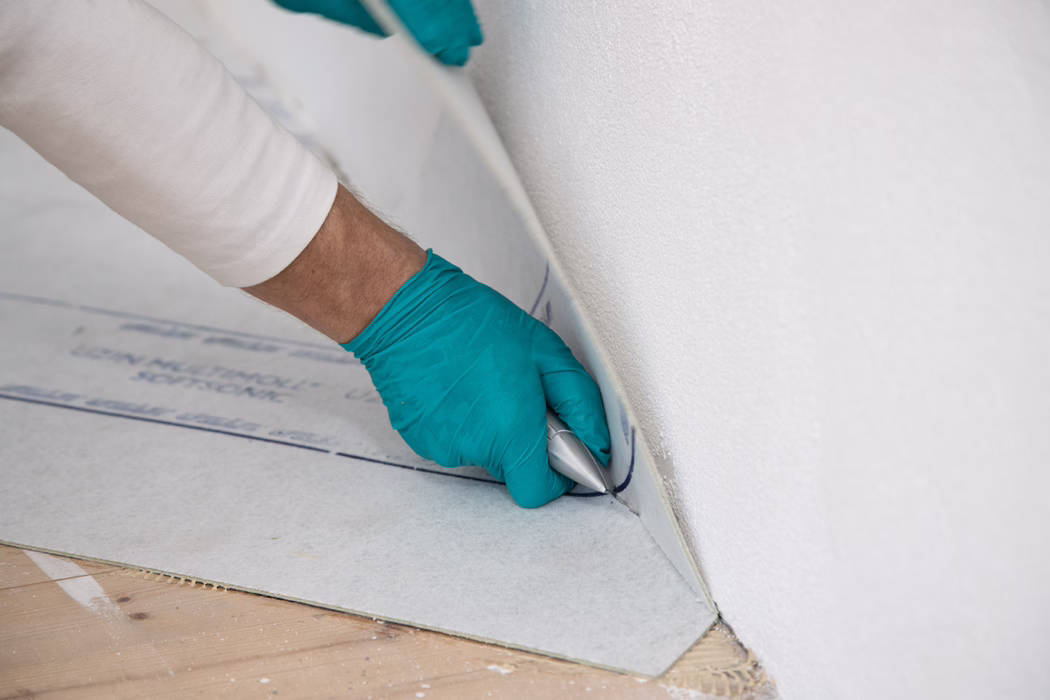 Gros plan d'une main gantée coupant ou ajustant un sous-plancher gris clair le long de la base d'un mur blanc avec un couteau utilitaire ; la main porte un gant jetable turquoise et le sous-plancher est replié pour suivre l'angle, laissant apparaître le sous-plancher en bois et quelques débris.