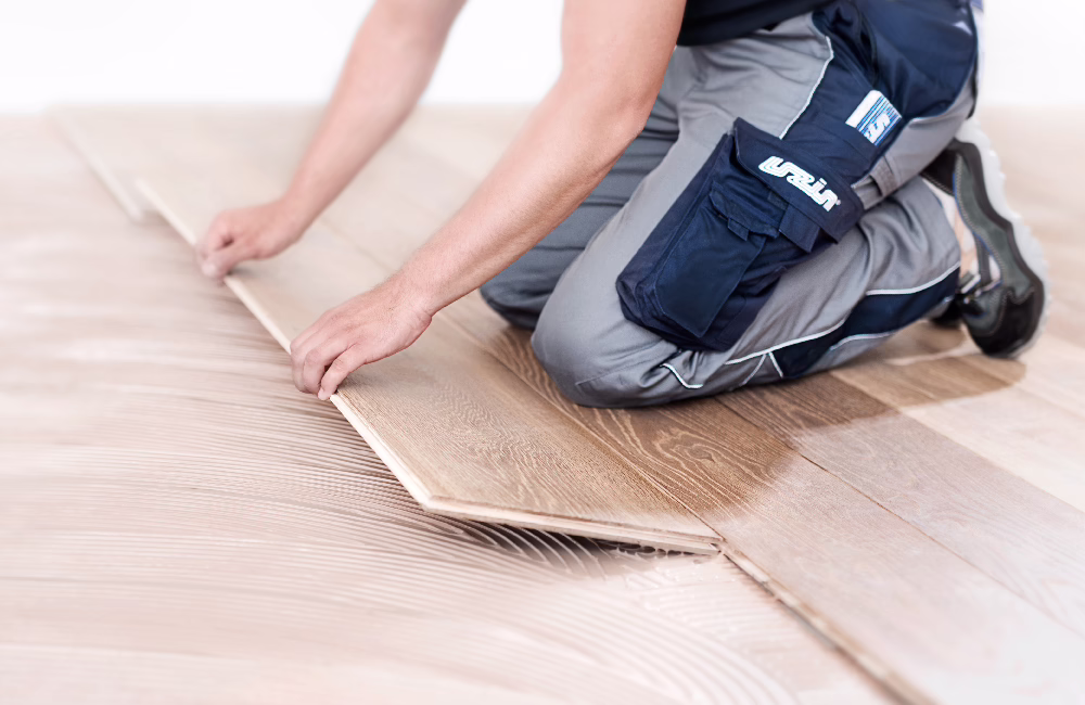 UZIN_Application Image_Parquet_2016-01 Un ouvrier agenouillé pose des lames de parquet stratifié chêne clair ; gros plan sur ses mains qui ajustent une lame sur de la colle, il porte un pantalon de travail gris et bleu marine avec poches et genouillères ; intérieur, éclairage uniforme, motif de lames décalées.