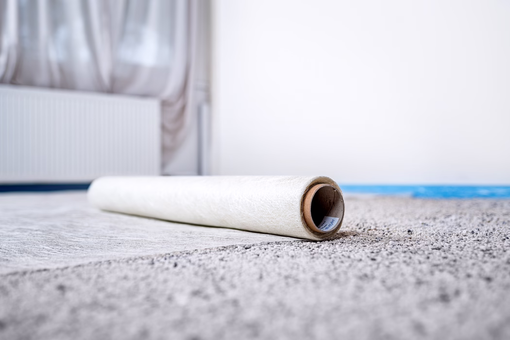 Gros plan d'un rouleau partiellement déployé de papier peint ou de sous-couche posé sur une moquette claire dans une pièce lumineuse et peu meublée. L'accent est mis sur l'extrémité en carton du rouleau au premier plan, tandis qu'en arrière-plan flou apparaissent en bokeh doux un radiateur et des voilages. Des tons neutres et une lumière naturelle créent une atmosphère calme et minimaliste, suggérant des travaux de rénovation ou une décoration intérieure en cours.