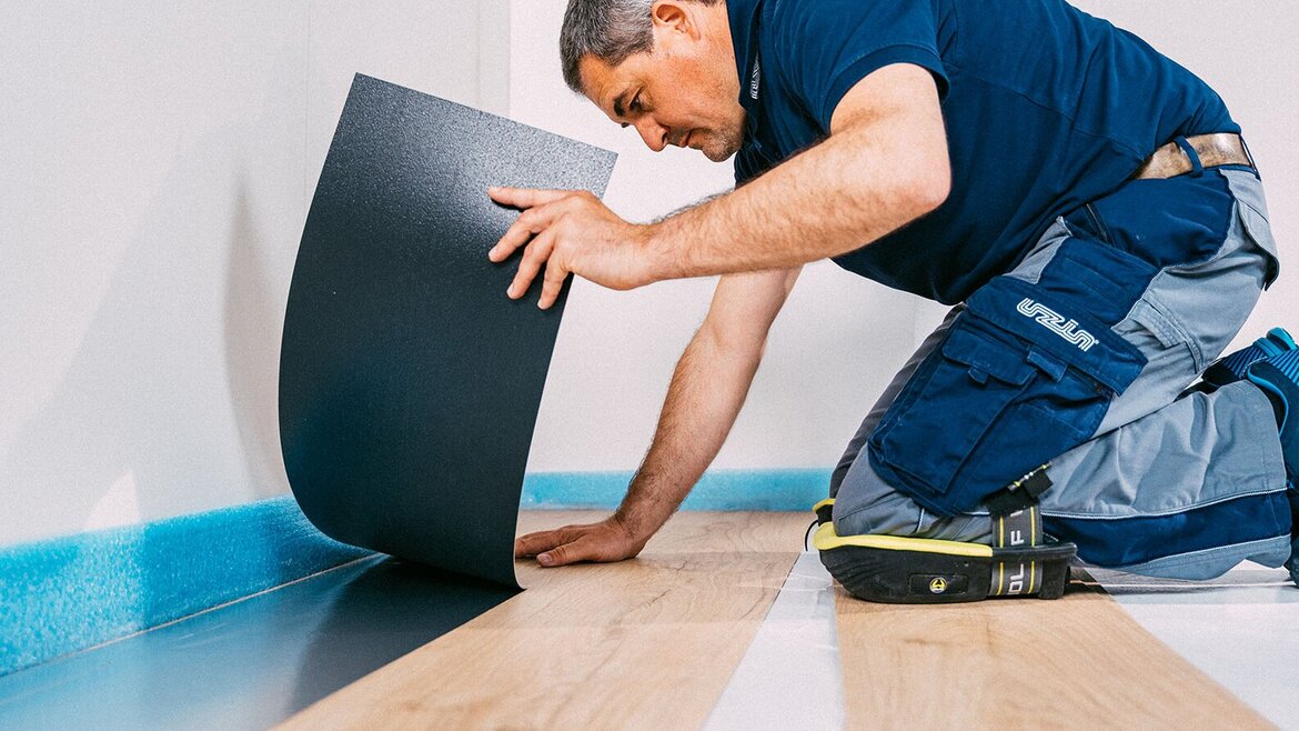 Un homme est agenouillé par terre, en train de poser un revêtement de sol en vinyle noir sur un parquet clair, dans une pièce aux murs blancs.