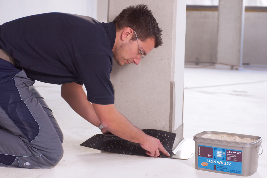 Un ouvrier en tenue de travail et portant des lunettes de protection est à genoux à l'intérieur d'un local, en train de poser et d'appliquer une membrane noire ou un revêtement adhésif au pied d'un mur ou d'une colonne claire. Il lisse le matériau du bout des doigts le long du bord du sol. À proximité se trouve un seau en plastique marqué 'UZIN WK 222' contenant de la colle. L'espace est un intérieur propre et inachevé avec un sol en béton ou en chape et peu d'éléments de fond.