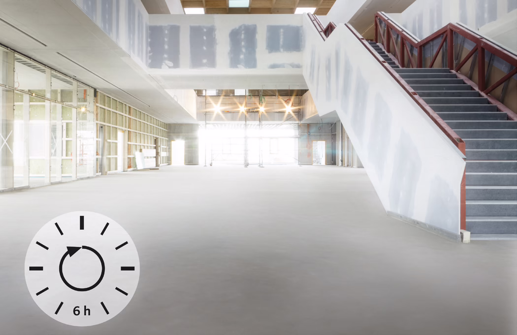 Intérieur spacieux et lumineux d'un bâtiment en construction ou en rénovation. Grande salle ouverte avec sol en béton lisse, cloisons sèches inachevées et ossature métallique apparente sur le mur gauche. Éclairages temporaires et échafaudages sont visibles dans l'entrée baignée de lumière à l'arrière ; un large escalier à droite, soutenu par une structure métallique et avec une rampe partiellement terminée, mène à l'étage supérieur. Dans le coin inférieur gauche, une icône circulaire avec une flèche dans le sens horaire et l'inscription « 6h » indique un temps de séchage ou de durcissement de six heures pour le sol ou le revêtement. L'ensemble dégage l'impression d'un chantier propre, organisé et très lumineux.