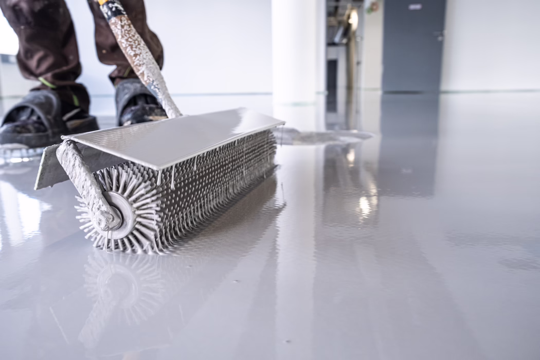 Gros plan d’un ouvrier appliquant un revêtement de sol autonivelant gris clair et brillant à l’aide d’un rouleau cranté d’aération. Le rouleau texturé répartit uniformément le produit liquide sur le sol intérieur, laissant une surface lisse et réfléchissante comparable à un miroir. Des gouttes de produit pendent du rouleau; on aperçoit les chaussures de protection de l’ouvrier et une porte floue en arrière-plan, suggérant un chantier en cours.