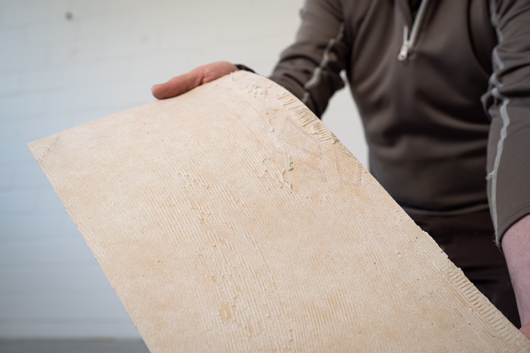 Gros plan d'une personne tenant une grande plaque rectangulaire beige présentant des stries d'adhésif texturé ; le torse et les mains de la personne sont partiellement visibles et elle porte un haut marron à manches longues. La plaque montre des rainures d'adhésif et de petites zones de plâtre ou de colle sèches.