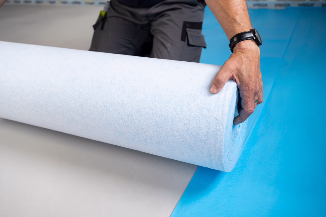 UZIN_Supplements & Accessories_PFB 160_blue_2024-06 (6) Gros plan sur la main d'un ouvrier déroulant un large rouleau d'isolant ou de sous-couche blanc et bleu pâle sur un sol bleu vif. La main droite porte une montre noire et une bague simple, saisissant le bord du rouleau ; un pantalon de travail gris avec poches et un outil clipsé sont partiellement visibles à l'arrière-plan. On distingue la texture douce et fibreuse du matériau ; la scène évoque un chantier intérieur ou une pose de revêtement de sol et la composition met en valeur la courbure du rouleau et le contraste de couleurs entre blanc, bleu pâle et bleu vif du sol.
