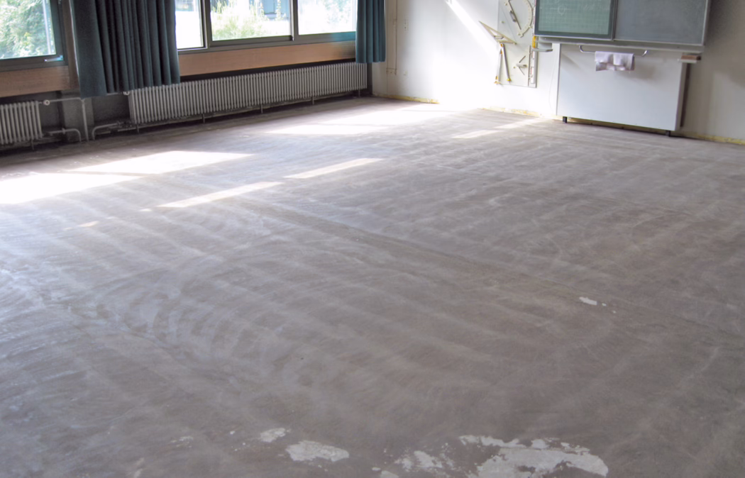 Une salle de classe lumineuse et vide dont le revêtement de sol a été retiré, révélant une sous-couche nue et poussiéreuse. De grandes fenêtres sur le côté gauche laissent entrer la lumière, formant des rectangles lumineux sur le sol ; des rideaux bleu-vert sont partiellement tirés et des radiateurs se trouvent sous les fenêtres. À l'avant à droite se trouvent un tableau blanc/tableau noir monté et quelques outils pédagogiques ; des traces d'adhésif ou d'enduit sont visibles sur le sol. La pièce est débarrassée du mobilier et semble en cours de rénovation ou de nettoyage en profondeur.