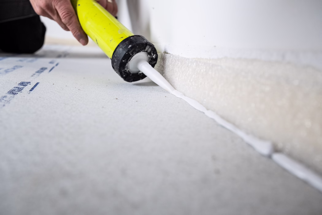 UZIN_Supplements & Accessories_Fondur HighTack_2024-03(46) Close-up of a person applying a continuous bead of white sealant from a yellow caulking gun along the joint between a wall and a foam insulation board on the floor. The plastic nozzle presses the fresh silicone into the gap where the textured foam meets the painted wall; part of the hand and the body of the caulking gun are visible on the left. The scene is lit with soft daylight, uses a shallow depth of field so the nozzle and sealant line are sharp while the foreground and background are blurred, and suggests a construction or renovation task focused on sealing and insulating edges.