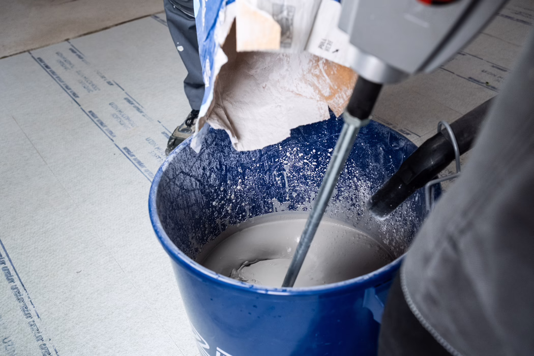 Gros plan d'un seau en plastique bleu posé sur le sol, rempli d'un mortier ou enduit gris en cours de mélange avec un mélangeur électrique manuel ; un sac en papier de poudre sèche est versé dans le seau, des éclaboussures couvrent les parois intérieures et les jambes et outils d'un ouvrier sont partiellement visibles.