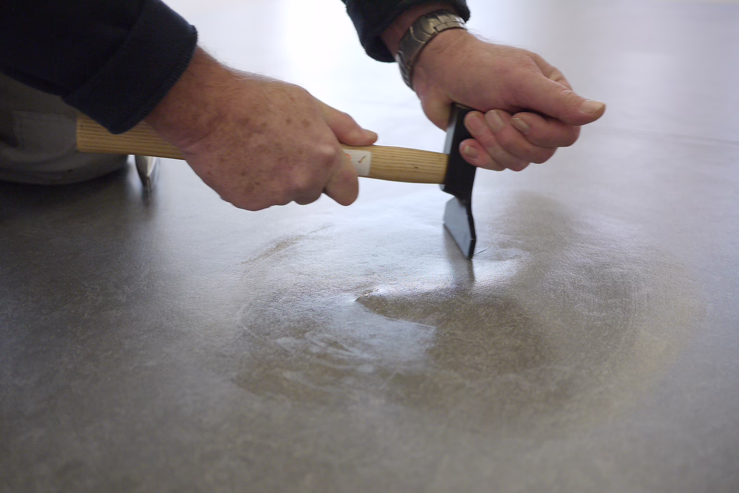 Gros plan sur les mains d'une personne agenouillée, lissant une surface de sol grise avec un outil semblable à un marteau et un outil à manche en bois ; la main droite tient une truelle métallique qu'elle enfonce dans la chape encore humide tandis que la main gauche maintient le manche en bois. Les mains présentent de petites taches de rousseur et une montre-bracelet ; l'arrière-plan est flou, l'accent est mis sur les outils et la texture du sol.