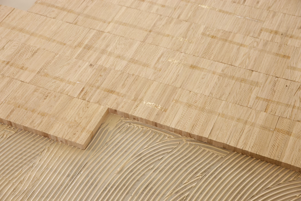 Gros plan d'un parquet en chêne clair partiellement posé. Des blocs de bois rectangulaires avec un grain visible sont collés sur le sous-plancher ; des traces de colle crantée apparaissent là où les panneaux suivants seront pressés. Une section du parquet est posée, montrant un alignement précis et des tons chauds et naturels du bois. L'image met en valeur la texture du chêne et le processus de pose.