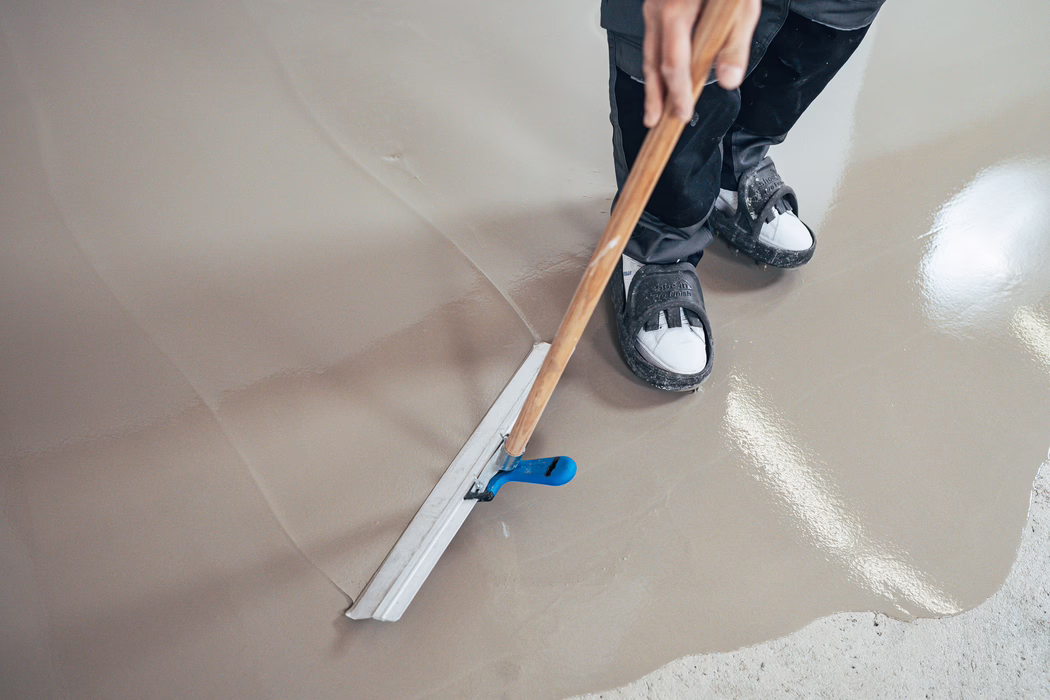 Gros plan d’un ouvrier lissant une chape liquide autonivelante fraîchement appliquée avec une taloche à long manche ; des chaussures de protection sont visibles et la surface apparaît brillante et humide.