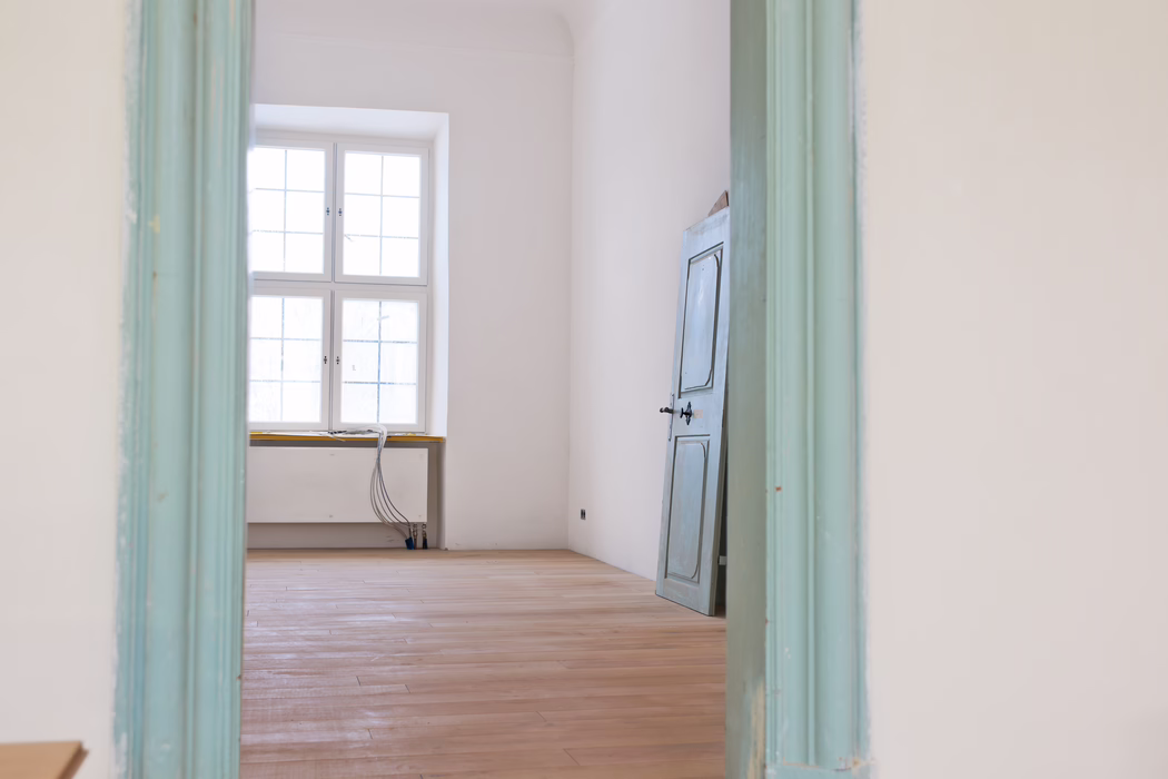 UZIN_Application_Image_MK 200_02_2018-10 Vue intérieure à travers un chambranle peint en turquoise pâle sur une pièce vide et baignée de lumière. Une grande fenêtre à quatre carreaux, centrée sur le mur du fond, inonde l'espace d'une lumière naturelle douce. Le sol est en lames de bois claires récemment poncées, les murs sont enduits de blanc et une porte en bois bleu gris légèrement patinée est appuyée contre le mur droit. Près de la fenêtre, des tuyaux de chauffage bas et un faisceau de câbles électriques apparents reposent sur le rebord et descendent au sol, suggérant des travaux de rénovation en cours. L'ambiance générale est calme et minimale, avec des tons pastel atténués et des lignes architecturales épurées.