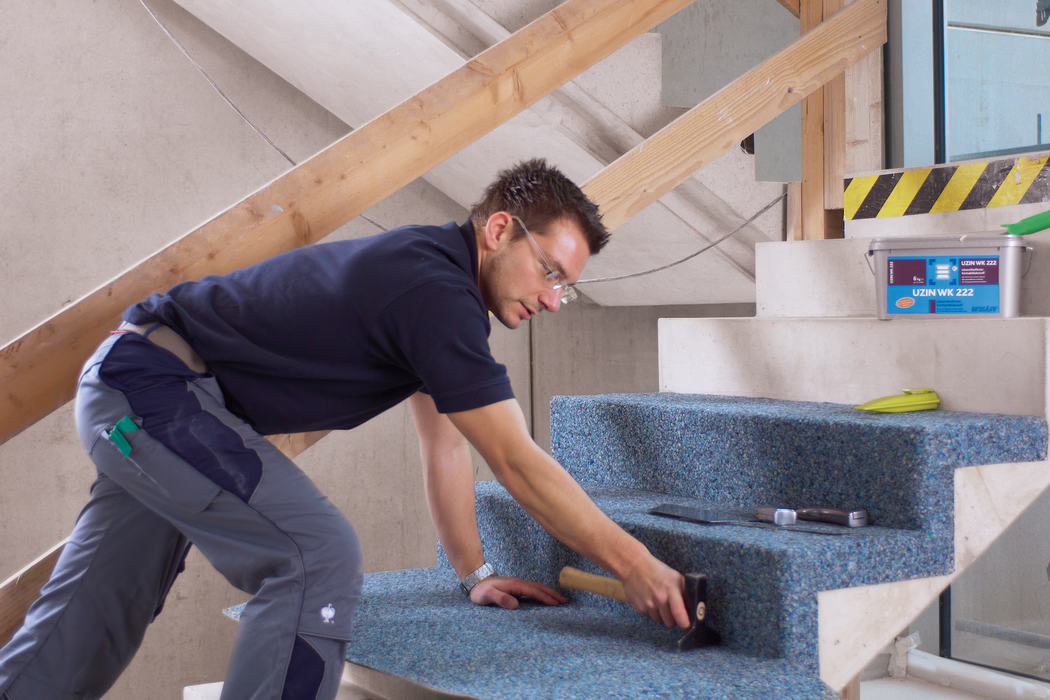 UZIN_Application_Image_WK 222_VA_4_ 2018-03 Un ouvrier pose une moquette bleue recyclée sur un escalier en béton non fini à l'intérieur d'un bâtiment en rénovation. Il est à genoux, penché en avant, porte des lunettes de protection, un polo bleu marine et un pantalon de travail gris, et utilise un maillet en caoutchouc pour bien fixer la moquette ; à proximité se trouvent une truelle, un mètre ruban et un seau en plastique marqué 'UZIN WK 222'. La scène est bien éclairée par la lumière naturelle, des planches de bois provisoires servant de rambarde sont visibles, l'accent est mis sur l'installation soignée et la texture de la moquette sur les marches.