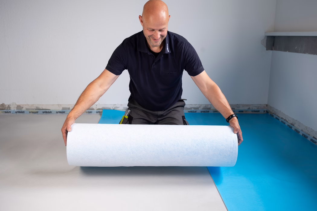 UZIN_Supplements & Accessories_PFB 160_blue_2024-06 (5) Un homme chauve d'âge moyen souriant, vêtu d'un polo sombre, déroule un rouleau blanc de sous-couche en posant un revêtement de sol sur une barrière vapeur bleue dans une pièce vide.