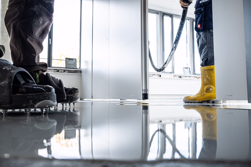 Deux ouvriers travaillent à niveler un sol fraîchement coulé et brillant à l'intérieur d'une pièce. L'ouvrier à gauche porte des chaussures à pointes pour marcher sur la chape autonivelante encore humide, tandis que l'ouvrier à droite, en bottes en caoutchouc jaunes, tient un tuyau qui déverse le mélange liquide. De grandes fenêtres laissent entrer une lumière naturelle vive et créent des reflets sur la surface brillante ; des outils et un radiateur sont visibles en arrière-plan.