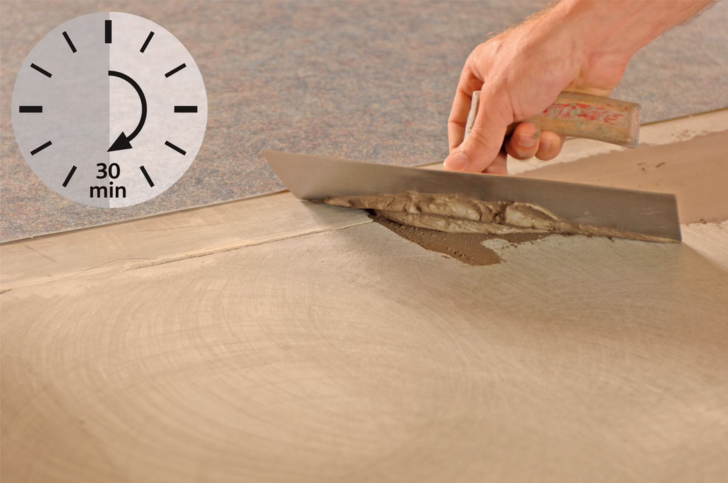 Une main tenant une taloche lisse un enduit de ragréage gris sur un sol en béton à côté d'un tapis ; une icône d'horloge indique « 30 min ».