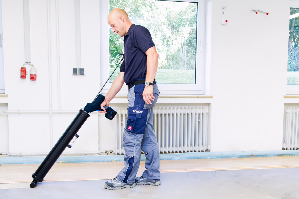 Un ouvrier d'entretien utilise un long appareil noir de nettoyage de sol à l'intérieur, debout devant une fenêtre et un radiateur, portant un polo bleu marine, un pantalon de travail gris et des chaussures de sécurité.