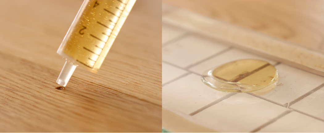 UZIN_Application_Image_RK 37_1_2018-03 Diptyque en gros plan montrant une colle jaunâtre distribuée à l'aide d'une seringue : le panneau de gauche montre une petite goutte qui se forme sur une surface en bois, le panneau de droite présente une goutte ronde du même adhésif sur des carreaux céramiques blancs avec un petit bouchon circulaire en bois partiellement enfoncé. Un éclairage chaud met en valeur le veinage du bois et les joints ; la faible profondeur de champ floute l'arrière-plan tandis que les graduations de la seringue et la texture de la colle restent visibles.