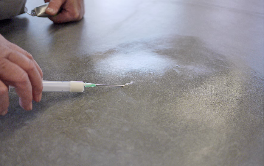 Gros plan sur deux mains réparant une petite déchirure dans une surface lisse en vinyle gris : une main tient une seringue avec une fine aiguille métallique insérée dans la déchirure, l'autre tient un petit outil métallique ou une cuillère. La surface présente une texture légèrement marbrée et des reflets doux provenant d'un éclairage au‑dessus. Une faible profondeur de champ concentre la mise au point sur la pointe de l'aiguille et la déchirure, tandis que l'arrière‑plan et les mains sont légèrement flous. Aucun visage n'est visible.