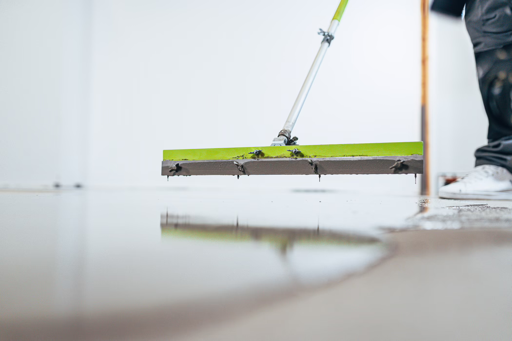 Close-up of a long-handled flooring squeegee spreading wet self-leveling compound across an indoor floor. The green metal blade is attached to an extendable pole; small clumps of compound cling to the screws and edges while droplets hang from the blade. The freshly leveled surface is glossy and reflects the tool. At the right edge a worker's lower leg and shoe are partially visible, suggesting a renovation or construction site inside a bright, minimal room.