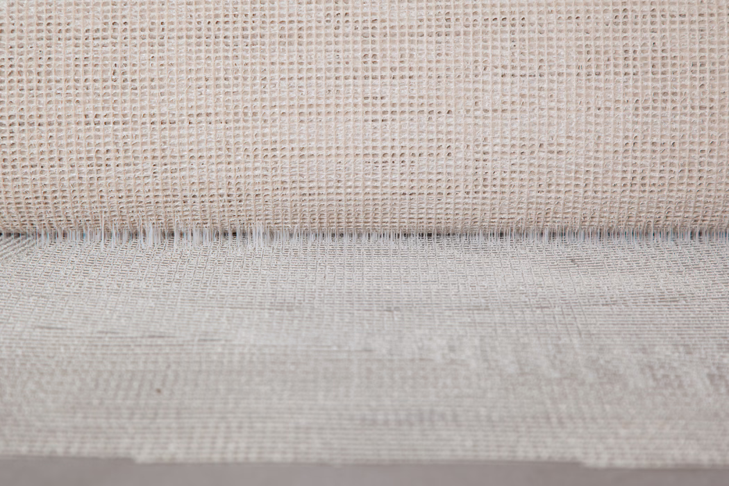 UZIN_Application Image_UZ57_2_6268_2018-11 Gros plan d'un tissu tissé beige neutre à motif de tissage ouvert en grille. La partie supérieure montre un bord roulé créant une ligne d'ombre fine; des fils irréguliers et des variations de fibres naturelles donnent un aspect tactile et rustique. Un éclairage doux et uniforme met en valeur la texture et la profondeur subtile.