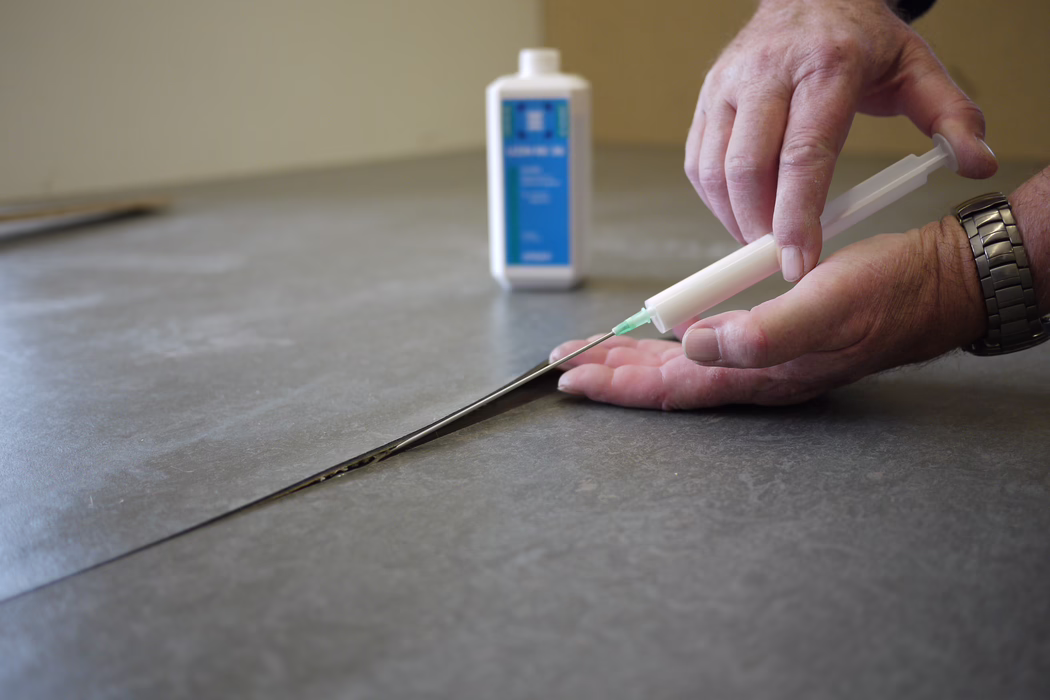 Gros plan sur des mains utilisant un applicateur en forme de seringue pour injecter de la colle ou un mastic dans une jointure d'un sol gris en vinyle ou stratifié, avec une petite bouteille floue en arrière-plan.