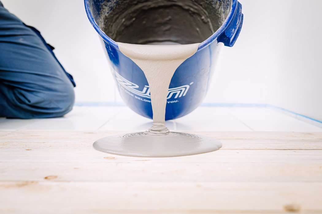 Gros plan d'un seau bleu versant un enduit de ragréage gris sur un sous-plancher en bois. Le mélange visqueux forme une flaque circulaire lisse sur le bois clair; à gauche, le genou d'un ouvrier en pantalon de travail foncé est visible et, à l'arrière-plan, un mur carrelé blanc et un ruban de peintre bleu.
