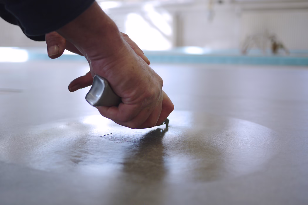 Gros plan d'une main tenant un petit grattoir ou spatule métallique pressé contre une surface horizontale lisse, créant un trait fin ou retirant du matériau ; lumière naturelle douce et faible profondeur de champ floutent l'arrière-plan.
