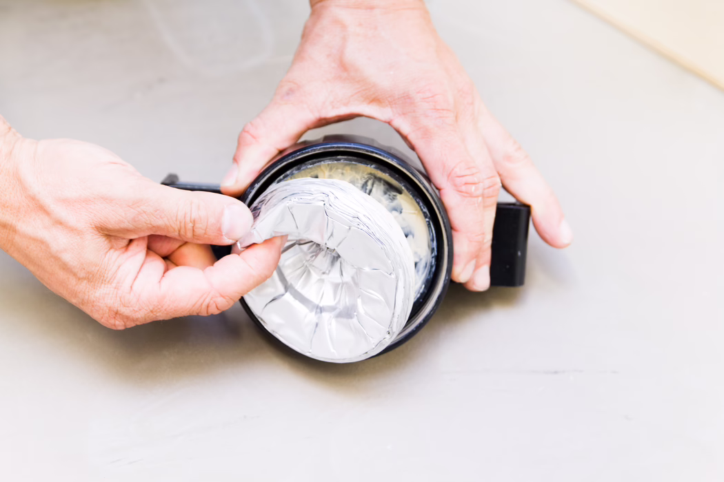 Gros plan de deux mains d'adulte tenant et assemblant un petit contenant rond noir tout en lissant ou en retirant une feuille d'aluminium froissée placée à l'intérieur. L'image est prise de dessus sur une surface gris pâle ; la feuille réfléchit la lumière avec des plis visibles et un anneau de colle ou de mastic borde le rebord intérieur. Les mains montrent une texture cutanée naturelle et de légères rides, ce qui suggère une réparation ou une opération d'étanchéité de type bricolage, avec des couleurs neutres et un faible profondeur de champ.