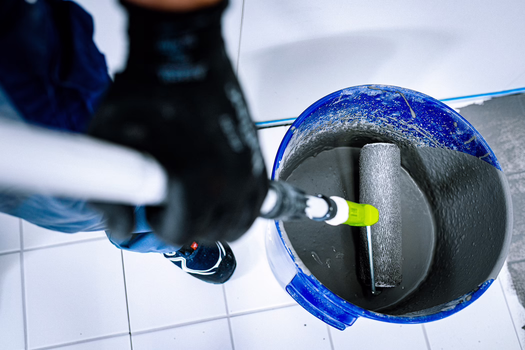 Vue plongeante d'un seau en plastique bleu rempli de peinture grise ou d'enduit, avec un rouleau à peinture reposant à l'intérieur. Une personne portant un gant foncé et un pantalon de travail bleu tient la poignée télescopique du rouleau ; un sol carrelé blanc et des chaussures de travail sont visibles. Le bord du seau est éclaboussé de peinture sèche et la housse du rouleau est uniformément couverte de matériau gris.