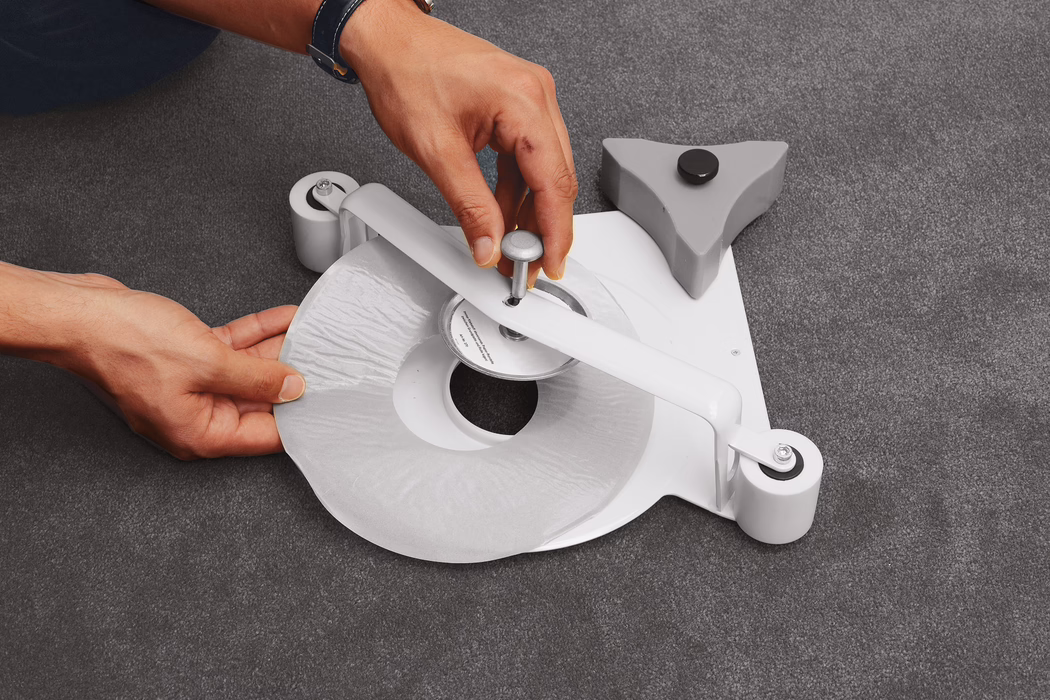 Gros plan sur des mains en train de poser ou de retirer un disque adhésif ou protecteur circulaire sur une base en plastique blanc équipée de deux roulettes et d'un axe central. Les mains ajustent soigneusement la pellicule circulaire au-dessus d'un moyeu métallique surélevé, tandis qu'un stabilisateur triangulaire gris avec une patinette noire est posé à proximité sur un tapis gris foncé. L'image met en valeur diverses textures — plastique blanc lisse, métal réfléchissant, film adhésif brillant et tapis à poils courts — avec des tons de peau chauds et une faible profondeur de champ qui concentre le regard sur les mains et l'assemblage central. On distingue des détails tels que le moyeu en forme de vis, les axes des roues et la surface légèrement froissée du disque protecteur.