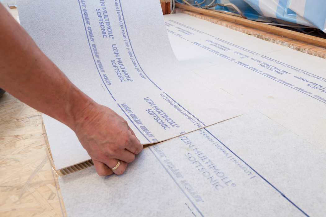 Gros plan d'une main posant un panneau isolant blanc avec l'inscription bleue 'UZIN MULTIMOLL SOFTSONIC' sur un sous-plancher en bois ; la main aligne les bords du panneau, on voit la construction en couches et la plaque OSB texturée en dessous.