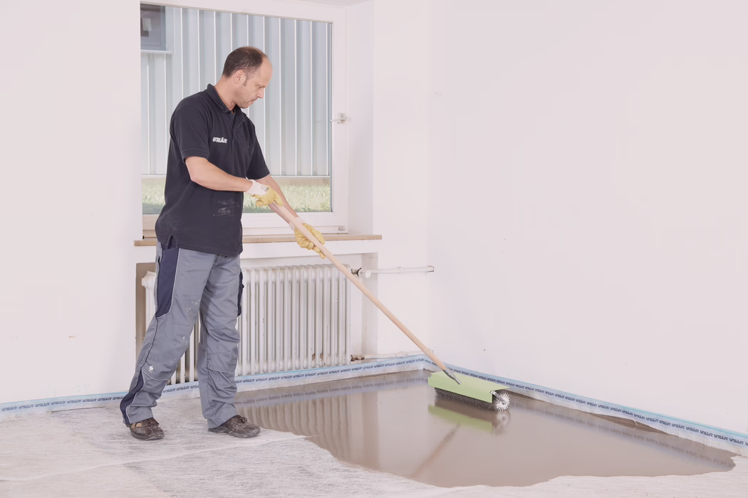 Un ouvrier, portant un pantalon de travail, un polo foncé et des gants jaunes, étale un enduit autonivelant avec un rouleau cranté à long manche dans une pièce vide et lumineuse. Il fait rouler l'outil le long du bord du sol près d'un radiateur blanc et d'une fenêtre, créant une surface lisse et brillante encore humide qui reflète la lumière. Les murs sont blancs, un ruban de protection est posé le long des plinthes et des taches de travail sont visibles sur les vêtements de l'ouvrier; la scène suggère une pose de sol professionnelle ou une rénovation.