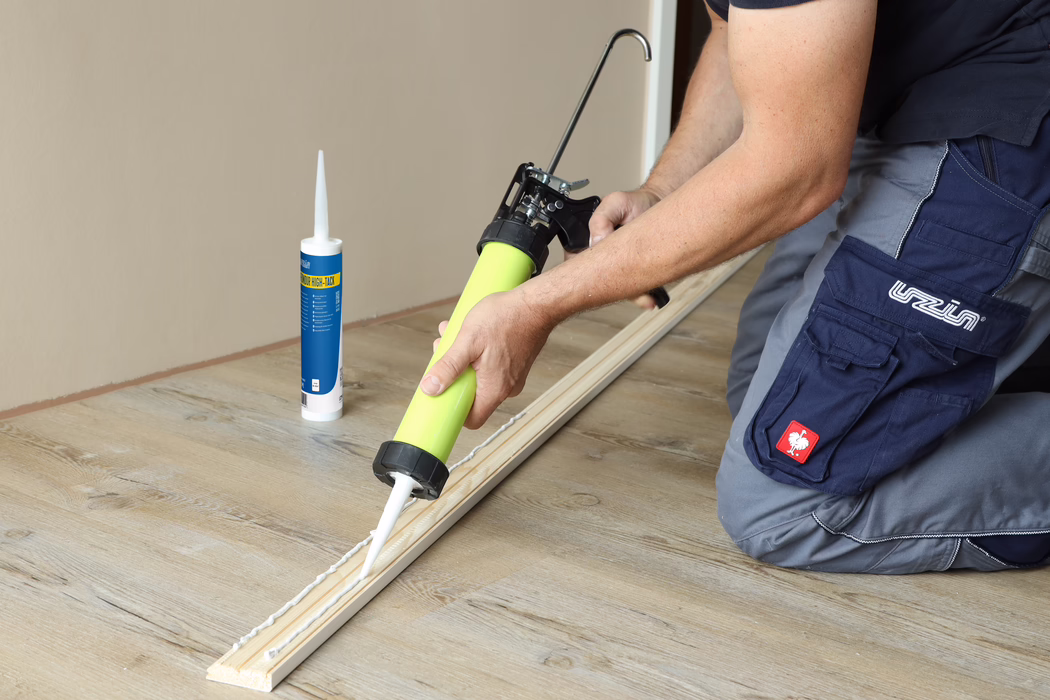 UZIN_Application Image_Fondur HighTack_104A6993_2022-03 Gros plan d'un ouvrier appliquant un mastic-colle blanc avec un pistolet à cartouche sur une baguette en bois posée sur un sol en bois clair. Une cartouche de colle est posée à proximité. L'ouvrier est à genoux et porte un pantalon de travail bleu ; l'image met l'accent sur les mains, le pistolet et le cordon de colle.
