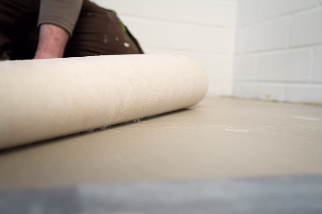 Gros plan d'une sous-couche de moquette beige déroulée sur un sol en béton dans une pièce aux murs en parpaings peints en blanc ; on distingue en arrière-plan le bras d'un ouvrier et son pantalon de travail marron.