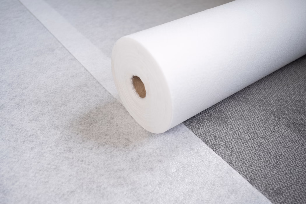 UZIN_Supplements & Accessories_PF120_Classic_2024-06 (15) Gros plan d'un grand rouleau de matériau blanc non tissé, ressemblant à du feutre, posé sur une moquette grise texturée. Le rouleau laisse apparaître un noyau en carton et une feuille partiellement déroulée qui s'étire sur le sol. Un éclairage doux et diffus fait ressortir la texture fibreuse fine, les variations subtiles de ton et les ombres légères ; la scène évoque une sous-couche de sol, une protection de tapis ou un matériau pour loisirs créatifs.