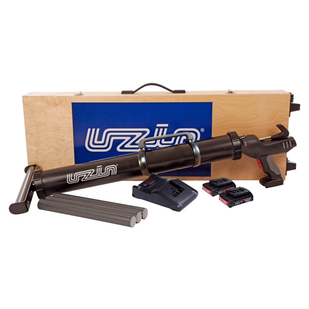 Mallette de transport en bois avec un pistolet à cartouche sans fil noir et ses accessoires : on distingue le dessus d'une mallette en contreplaqué clair avec une poignée noire et un panneau logo bleu, derrière un long pistolet à cartouche noir alimenté par batterie. Devant la mallette se trouvent trois cartouches grises, une base de chargeur et trois accus lithium-ion 18V. Le pistolet présente une poignée pistolet avec des accents gris et rouges, des colliers métalliques autour du canon de cartouche et une gâchette. Composition de studio sur fond blanc.